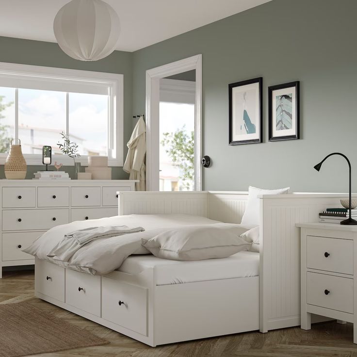 a white bed sitting in a bedroom next to a dresser and window with pictures on the wall