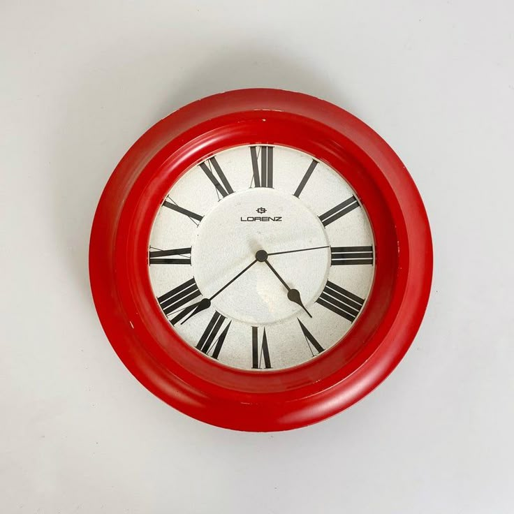 a red clock with roman numerals on the face is hanging on a white wall