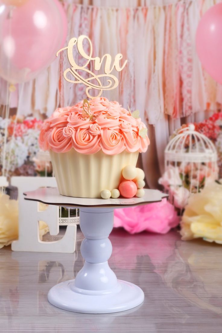 a cupcake with pink frosting on top sitting on a cake stand in front of balloons