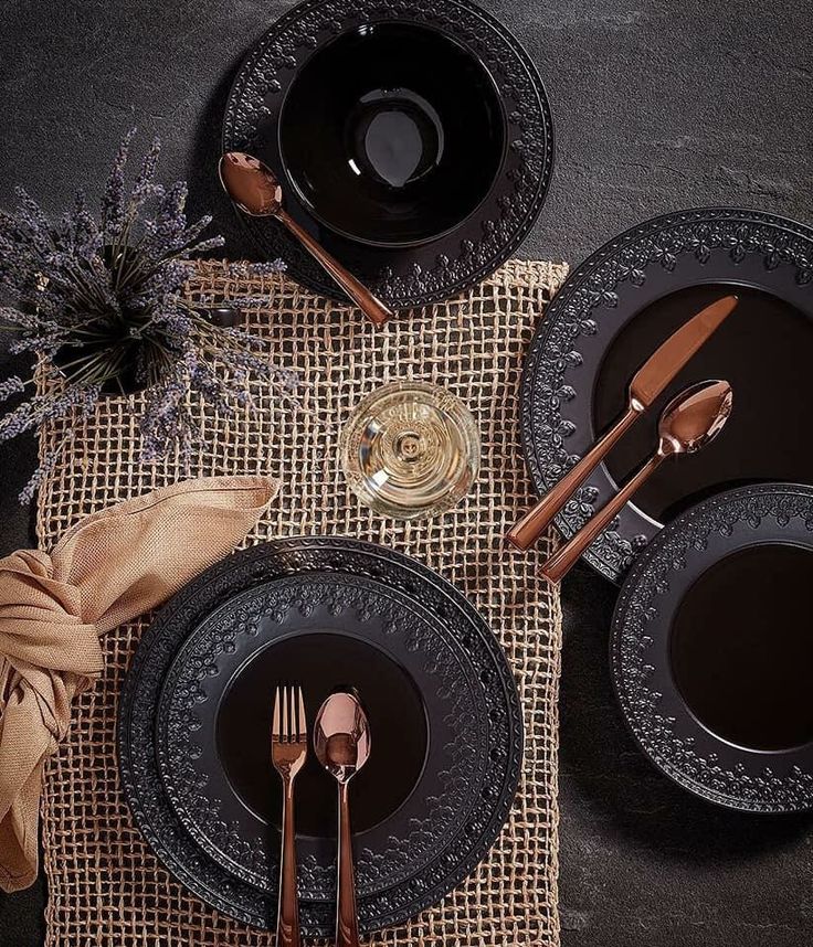 a table setting with black plates, silverware and gold utensils on a burlap placemat