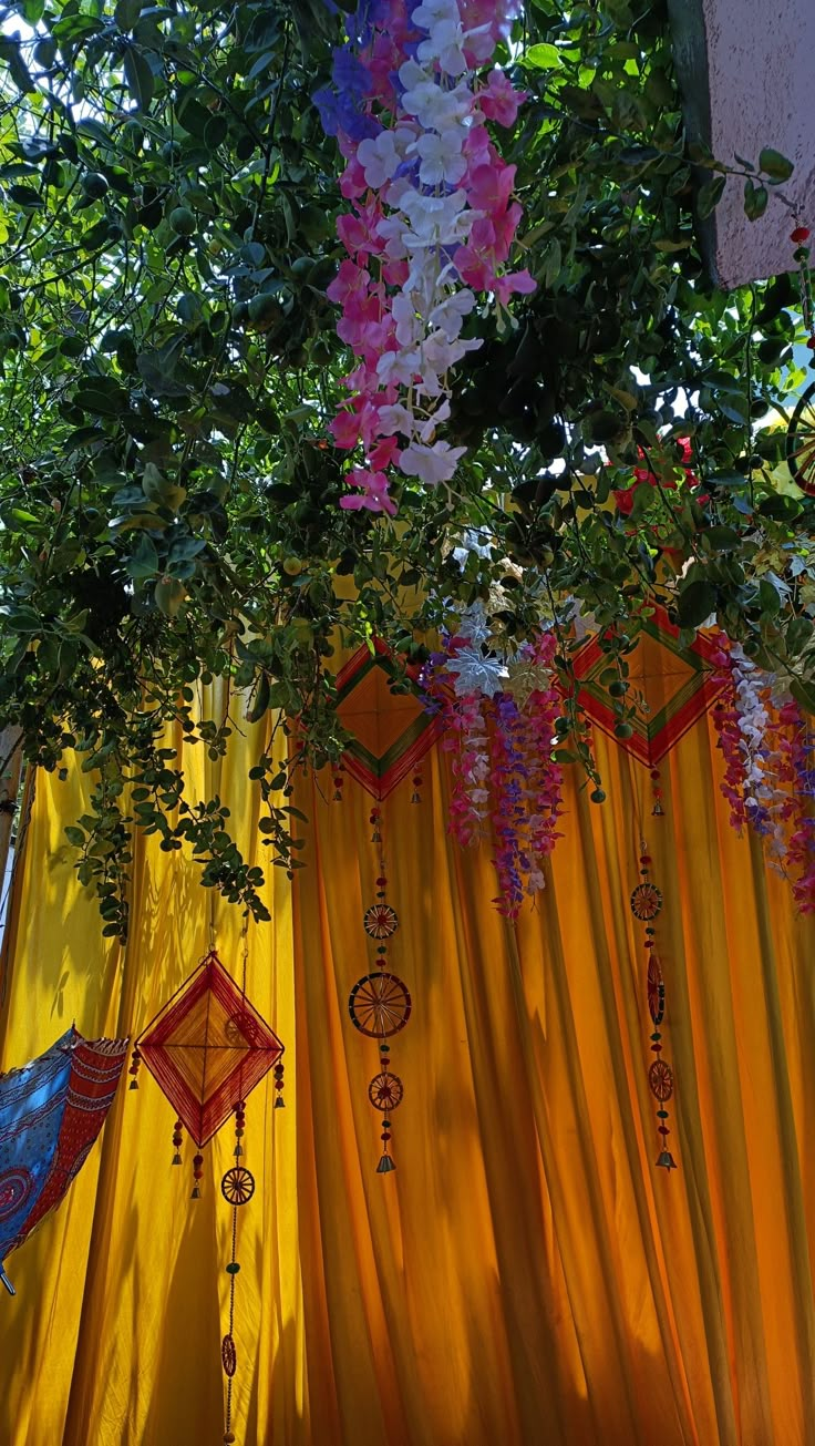 colorful decorations hanging from the ceiling in front of a yellow drape and tree branches