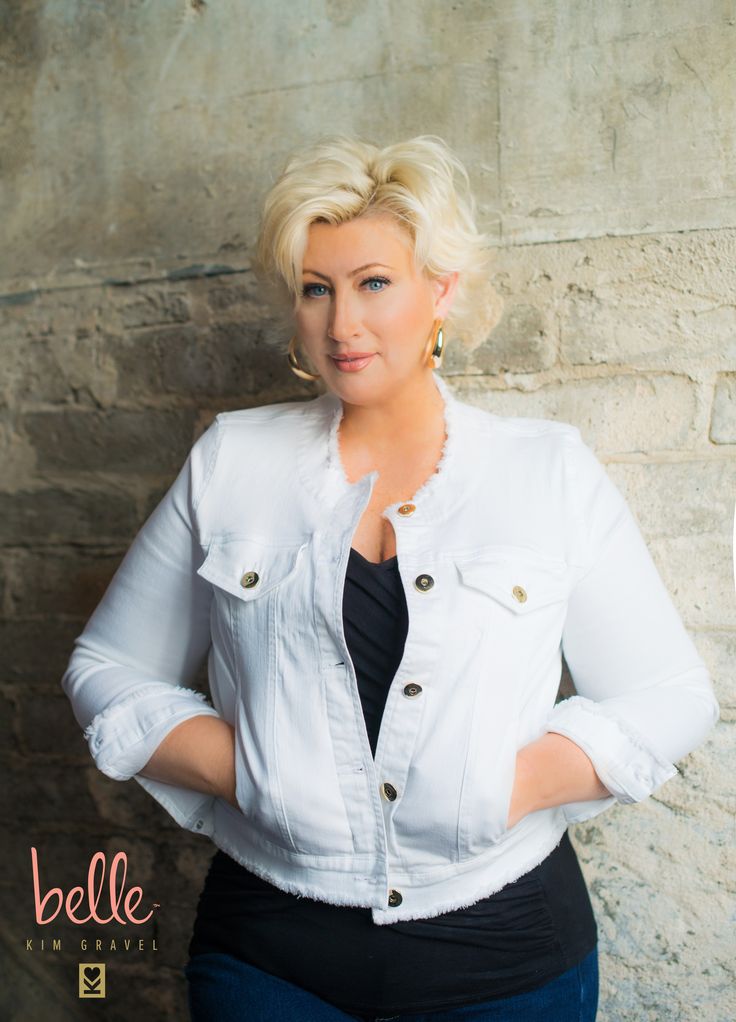 a woman with blonde hair wearing a white jacket and black top standing in front of a stone wall