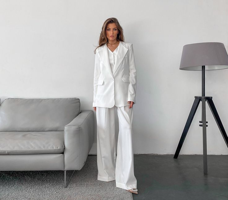 a woman standing in front of a couch wearing a white suit and matching pants with heels