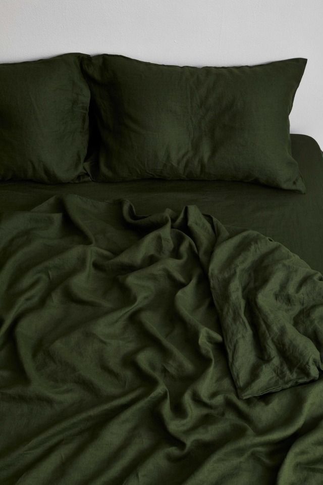 an unmade bed with green sheets and pillows on top of it, in front of a white wall