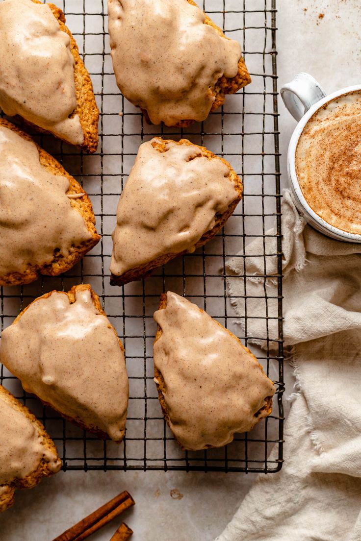 cinnamon buns with frosting and cinnamon sticks on a cooling rack next to a cup of coffee