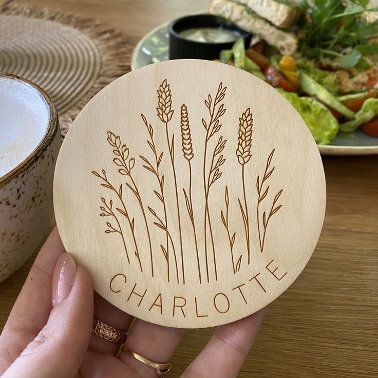 a person holding up a personalized wooden coaster with the name charlotte in front of a plate of salad