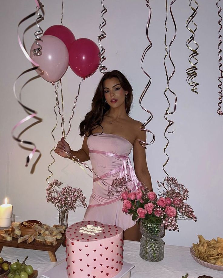 a woman in a pink dress standing next to a table with cake and balloons on it