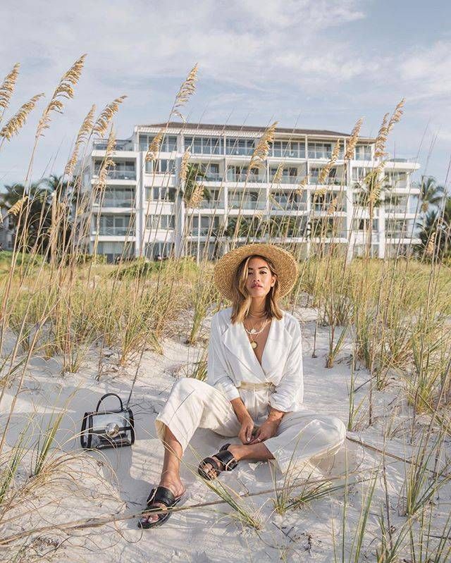 a woman sitting in the sand with a hat on her head and purse next to some tall grass