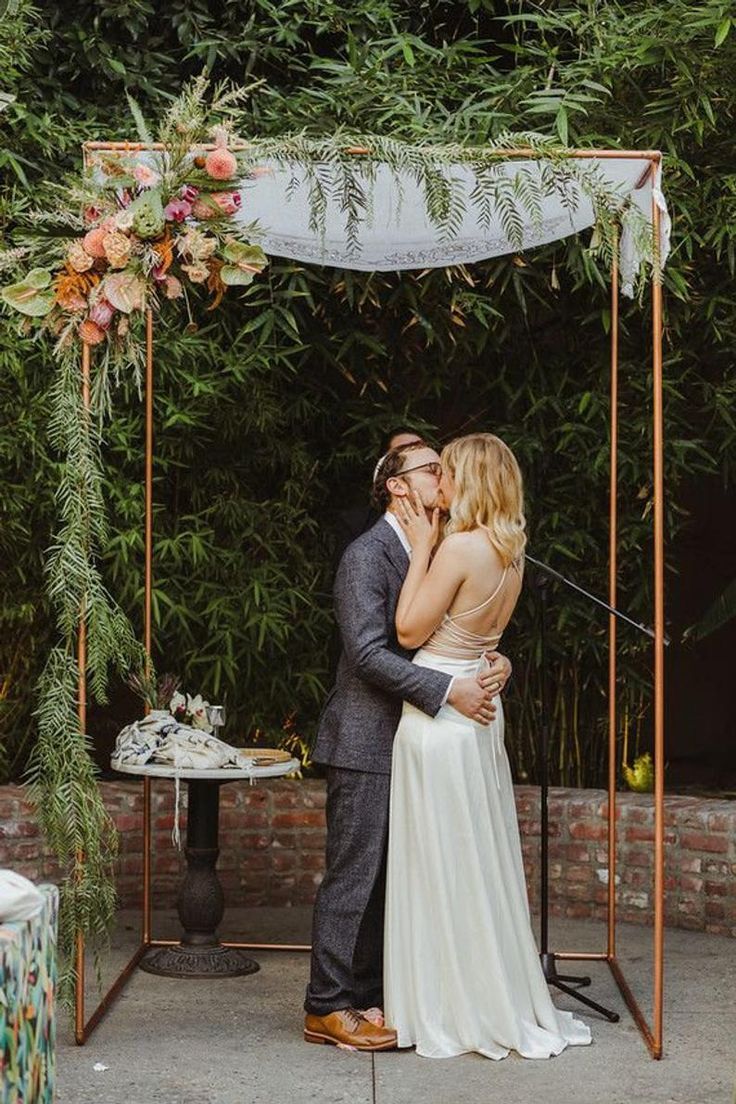 a bride and groom kissing under an outdoor wedding ceremony arch with greenery on the side