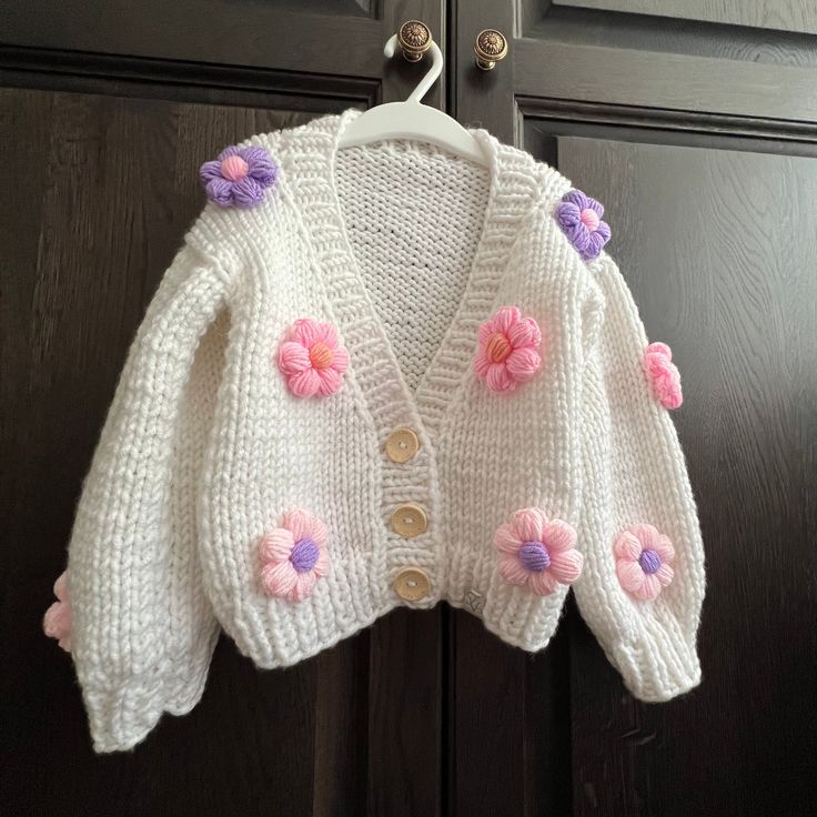 a white sweater with pink and purple flowers on it hanging from a wooden door handle