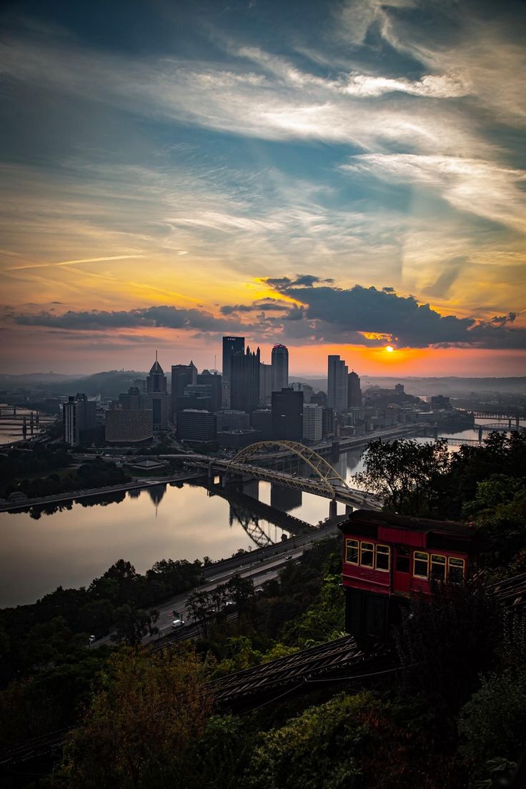 a red train traveling over a bridge next to a large city under a cloudy sky