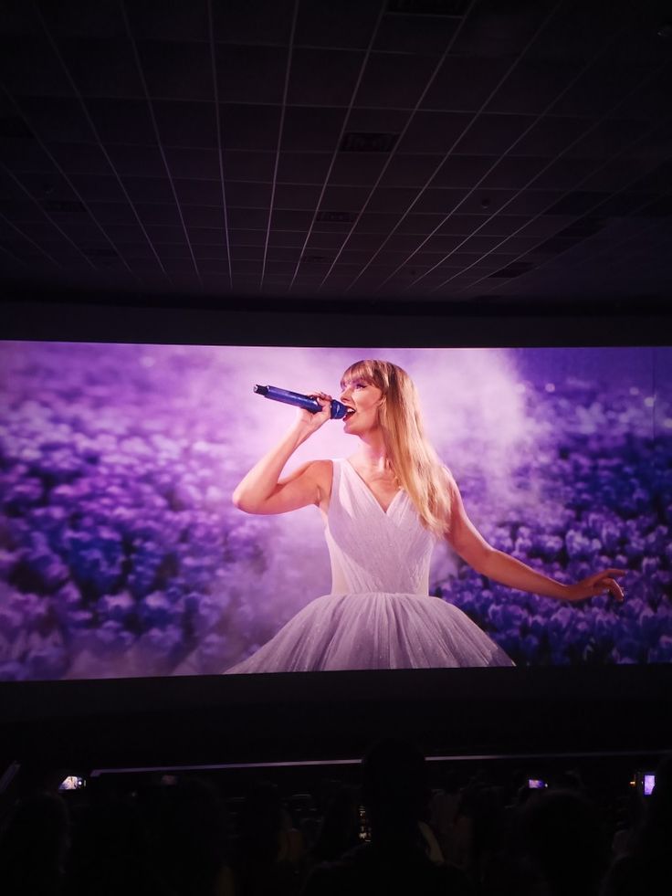 a woman in white dress holding a microphone on stage with large screen behind her and purple flowers