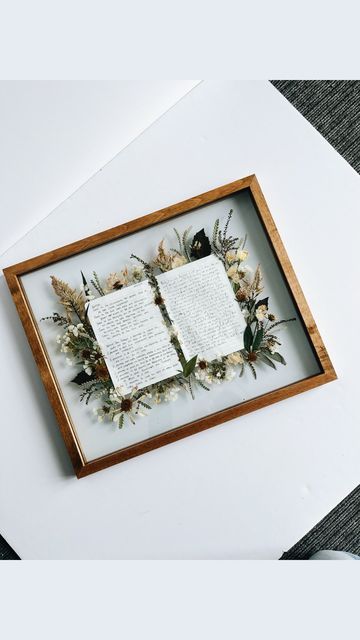 an open book with paper and flowers on it in a wooden frame hanging on the wall