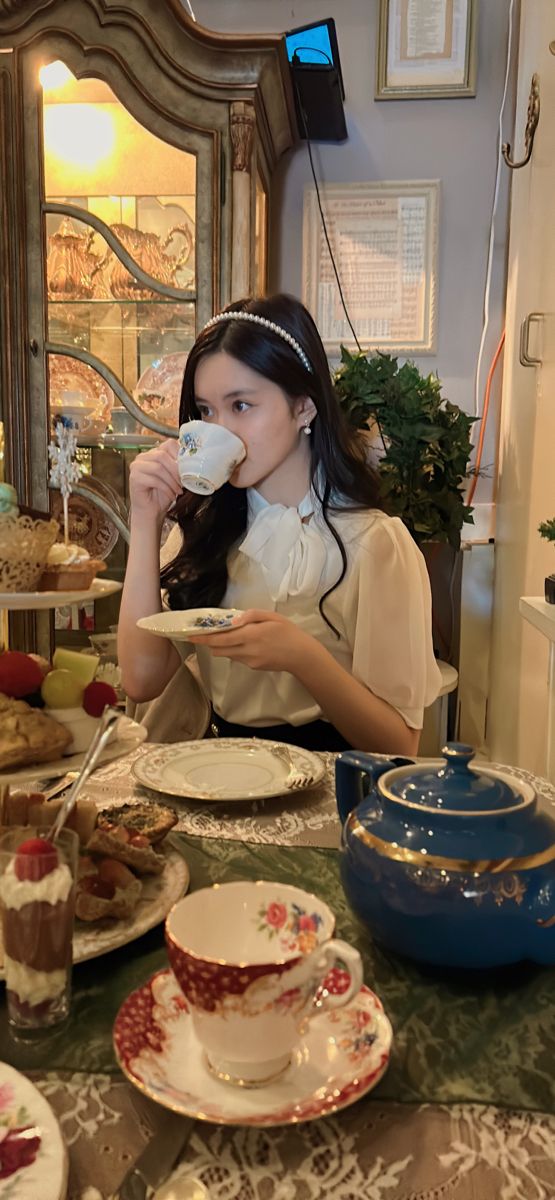 a woman sitting at a table drinking from a cup and saucer while holding a plate with desserts on it