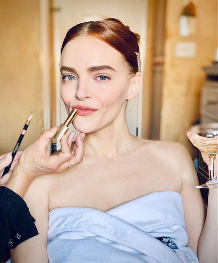 a woman in a blue towel holding a brush and lipstick on her cheek while looking at the camera