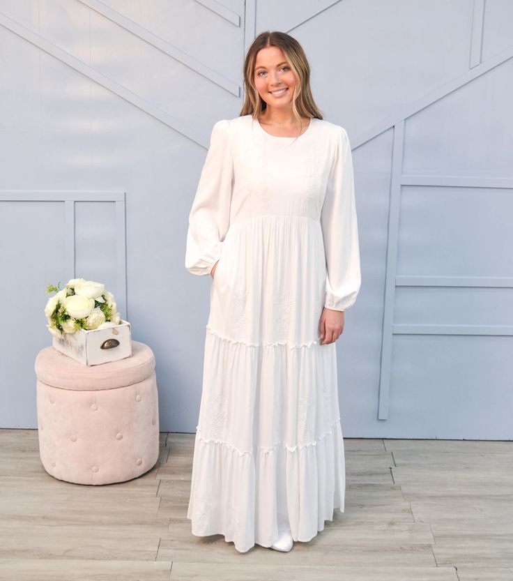 a woman standing in front of a wall wearing a white dress and smiling at the camera