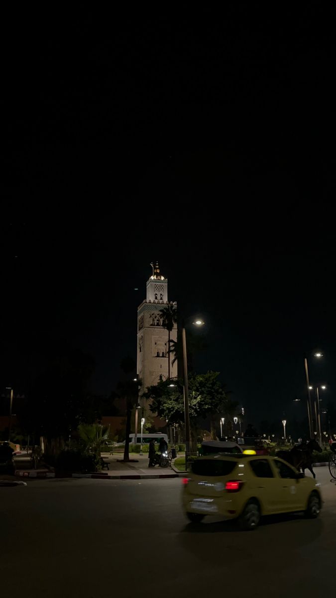 a yellow car is driving down the street at night with a clock tower in the background