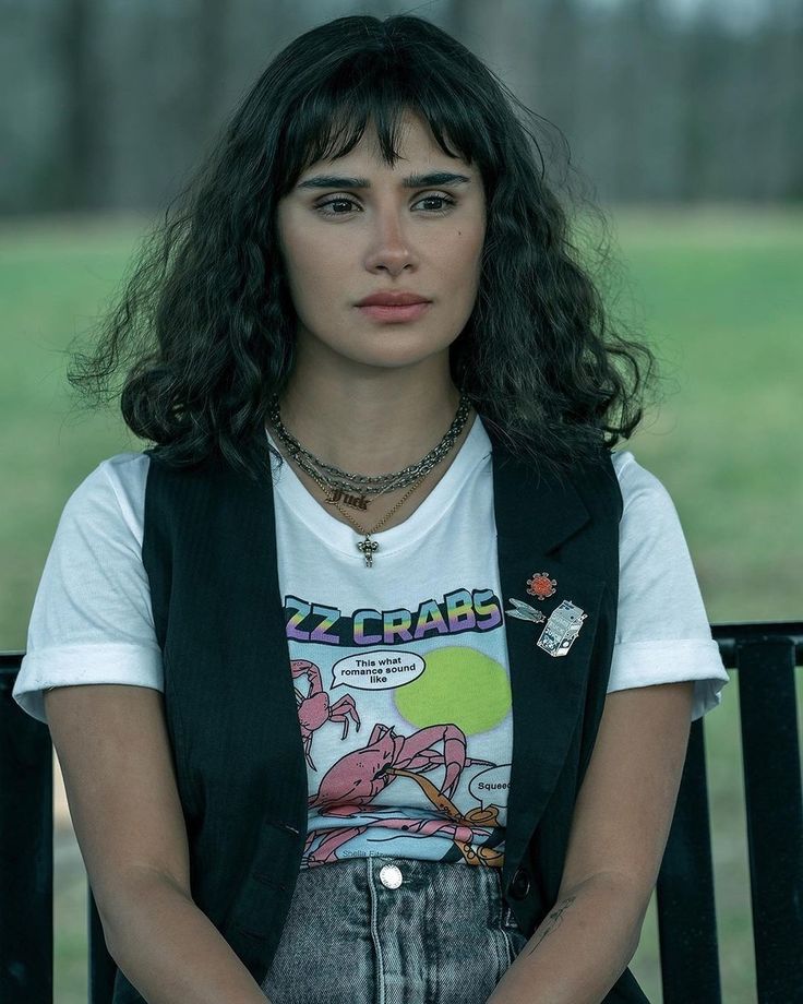 a woman sitting on a bench with her hands in her pockets and looking at the camera