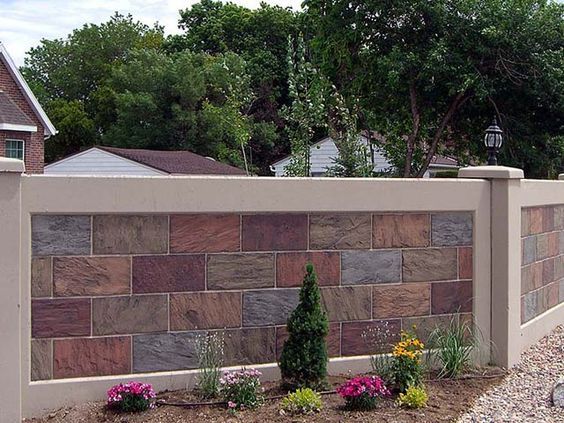 a brick fence with flowers and bushes in the foreground, along with a house on the other side