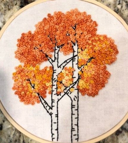 an embroidery project with orange and yellow trees in the middle on a white hoop hanging from a marble countertop