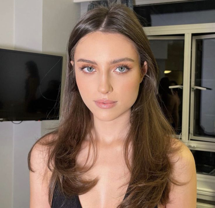 a woman with long brown hair wearing a black top and posing for the camera in front of a window