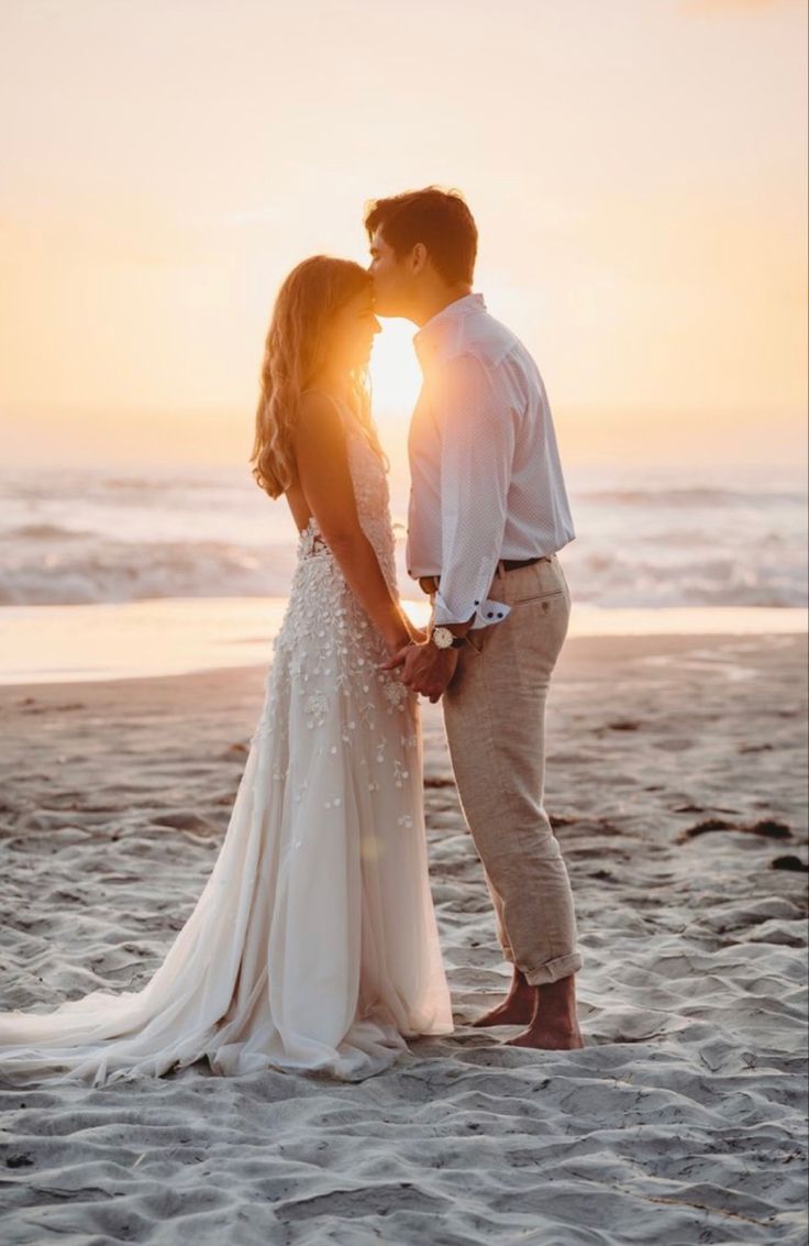 a bride and groom kissing on the beach at sunset