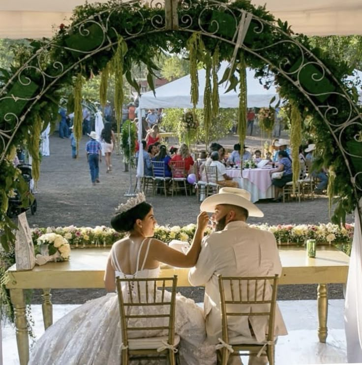 a man and woman sitting at a table in front of an arch decorated with greenery