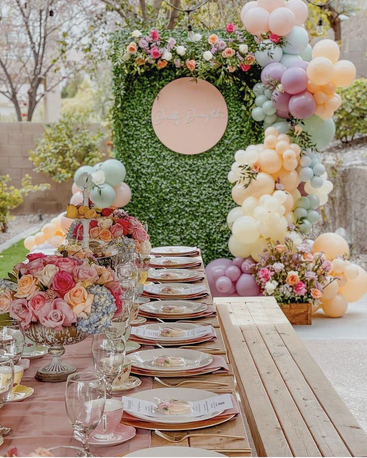 a table set with plates, glasses and flowers in front of an outdoor wall decoration