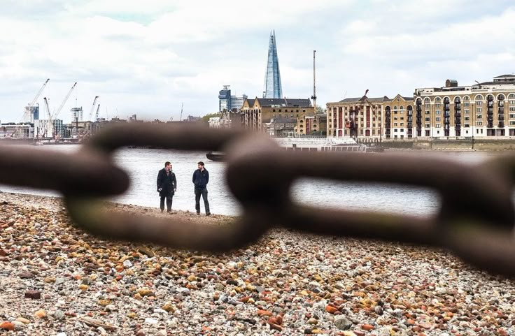 two people standing in the water behind a chain link fence with buildings in the background