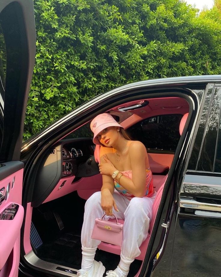 a woman sitting in the driver's seat of a pink car with her hand on her chest