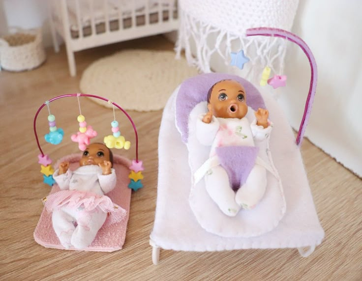 two small dolls sitting on top of a white blanket next to a toy baby in a swing