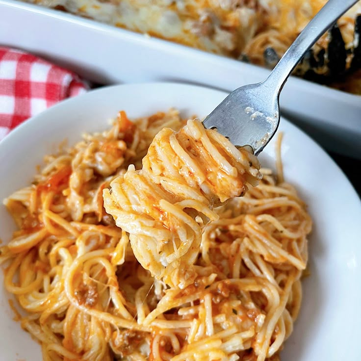 a fork full of spaghetti on a white plate next to a casserole dish