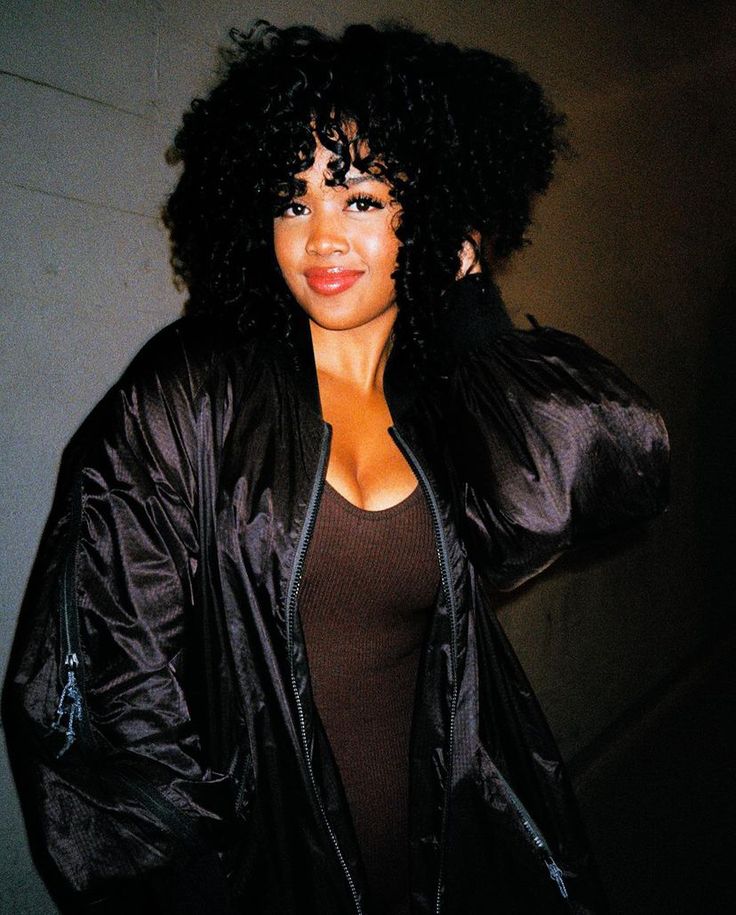 a woman with curly hair wearing a black jacket and posing for the camera in front of a wall