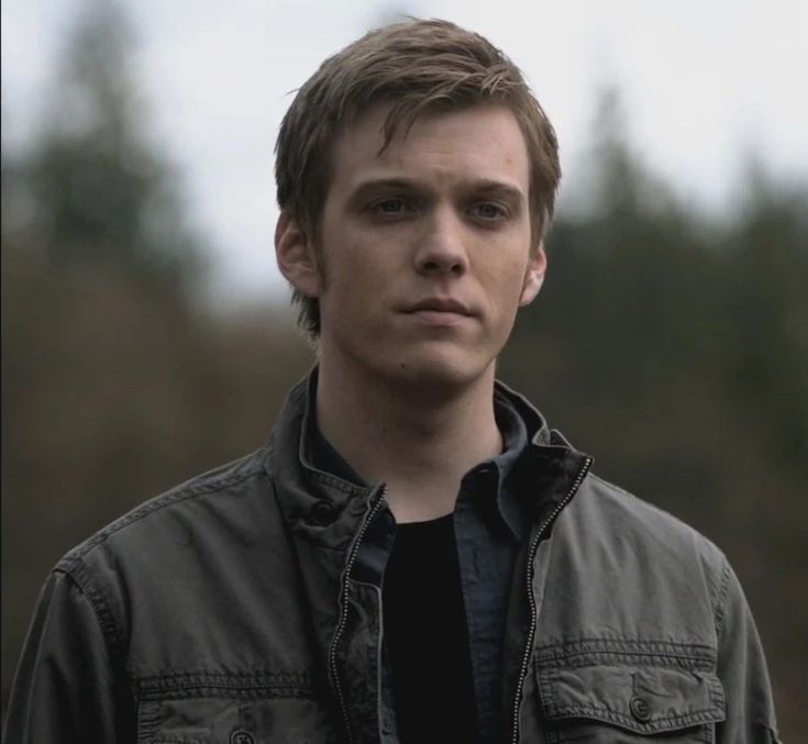 a young man in a jacket looks at the camera while standing outside with trees in the background