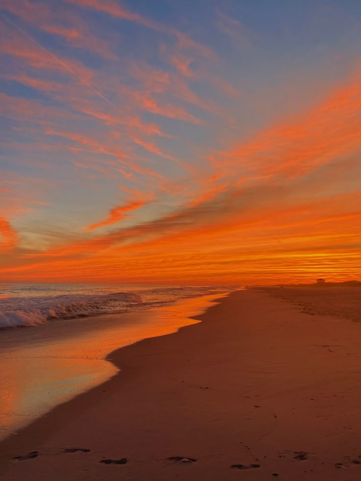 an orange and blue sunset over the ocean with footprints in the sand on the beach