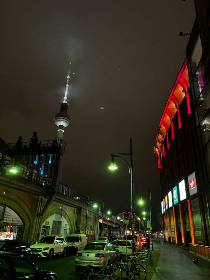 a city street at night with cars parked on the side and tall buildings in the background