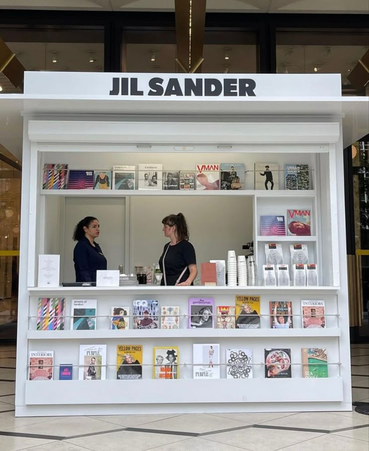 two women are standing in front of a book store with shelves full of books and magazines