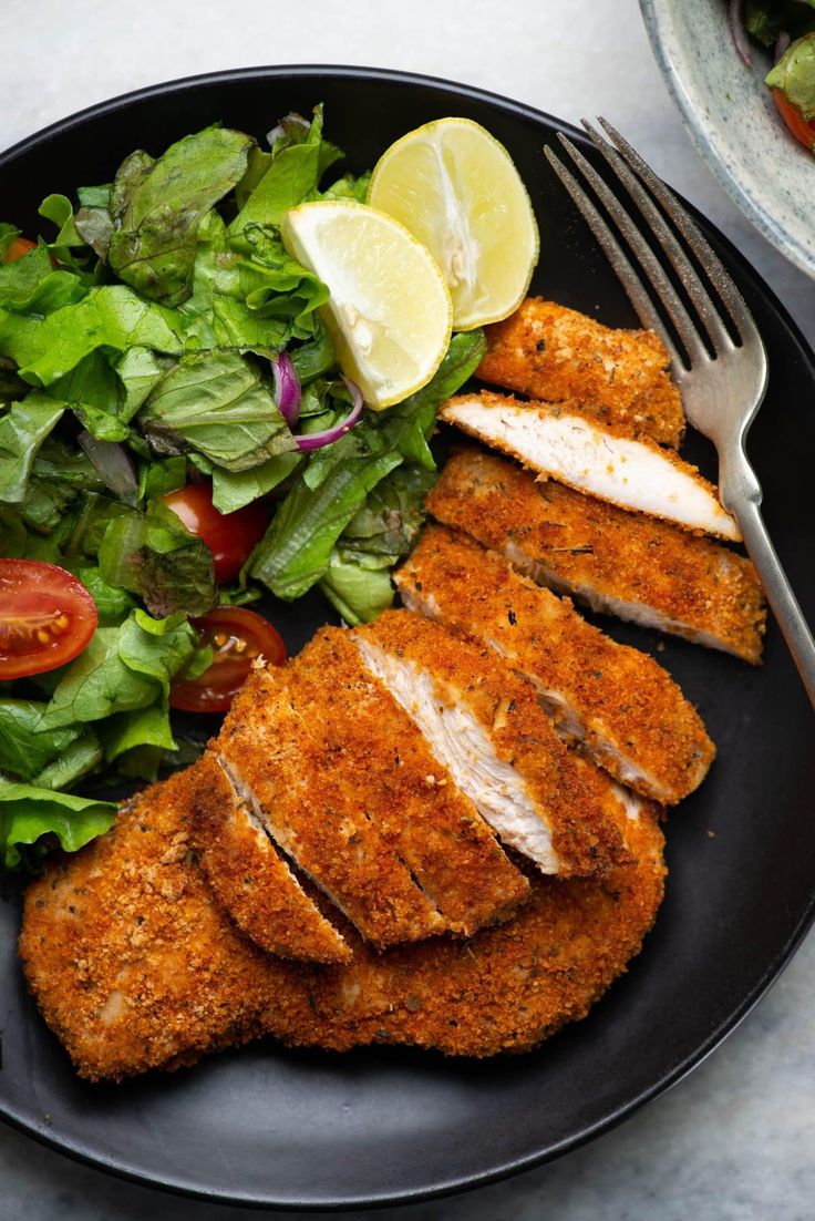 a black plate topped with chicken, lettuce and tomato salad next to a fork