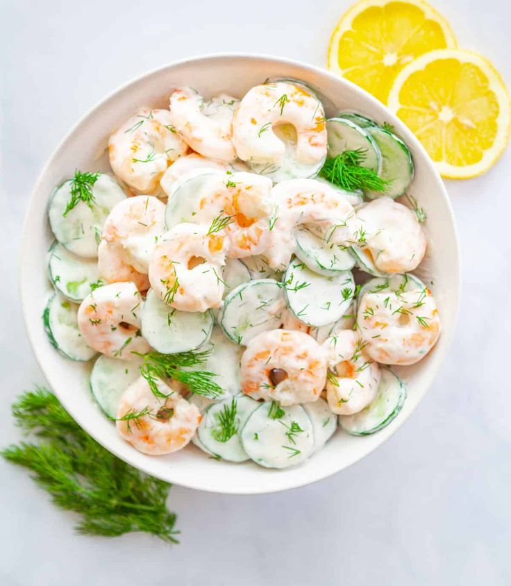 a bowl filled with shrimp and cucumbers next to lemon wedges on a white surface