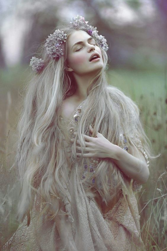 a woman with long hair and flowers in her hair is standing in the grass, looking up