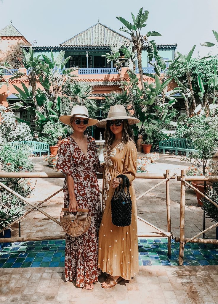 two women standing next to each other in front of a building with trees and plants