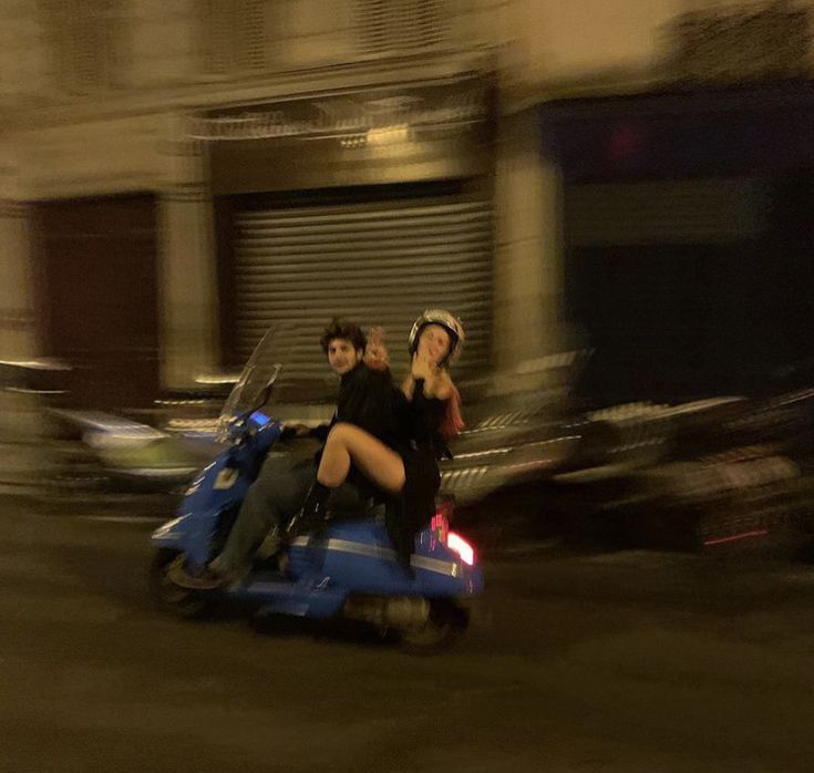 two people riding on the back of a blue scooter in front of a building