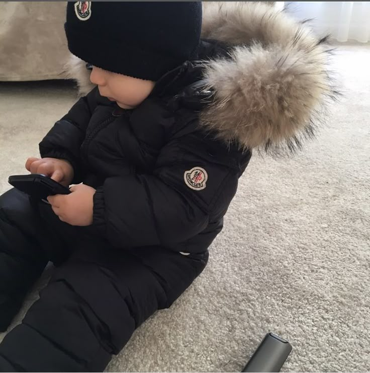 a small child sitting on the floor playing with a cell phone and wearing a winter coat