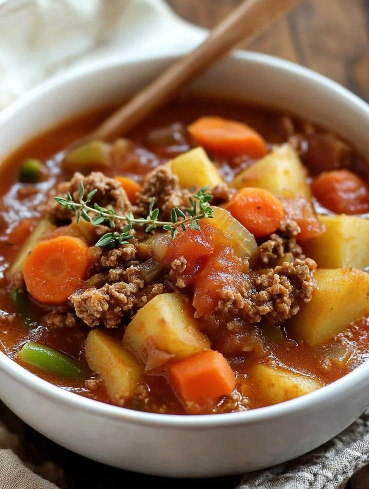 a white bowl filled with stew and carrots next to a wooden spoon on top of a napkin