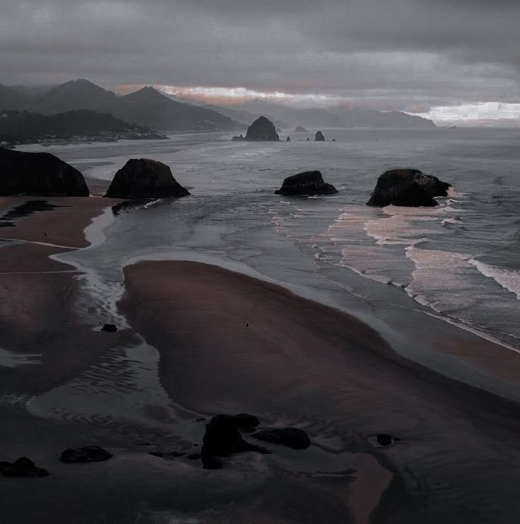 an ocean beach with rocks in the water and mountains in the distance on a cloudy day