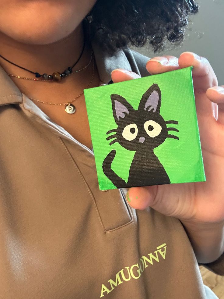 a woman holding up a small painting of a black cat on a green background with the words amoung