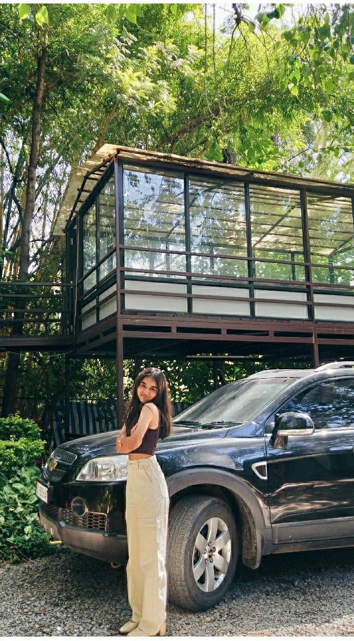 a woman standing next to a black car in front of a house with glass walls