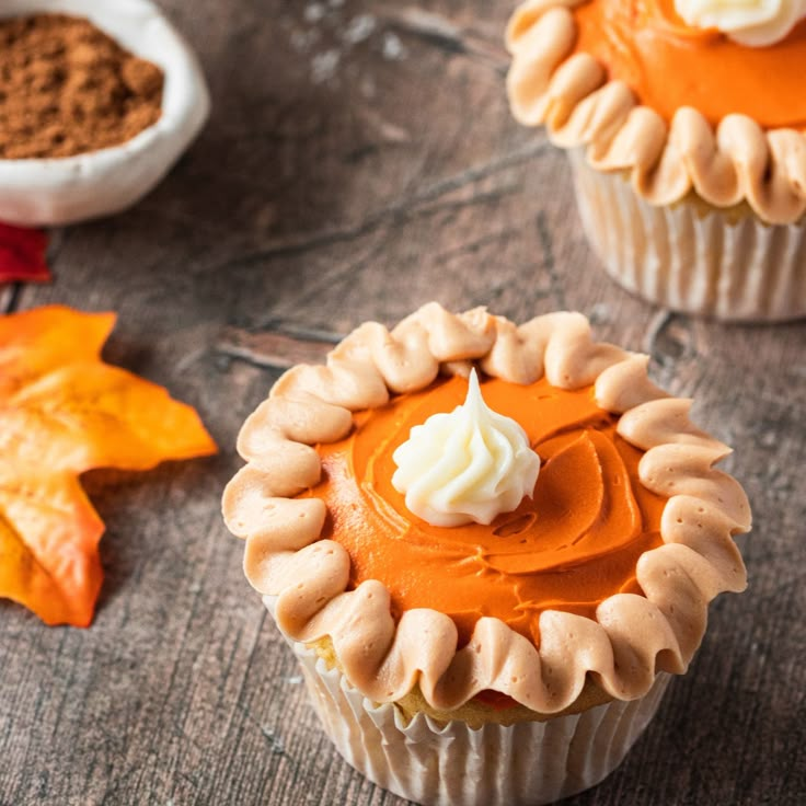 two cupcakes with orange frosting and white icing on top, next to some autumn leaves