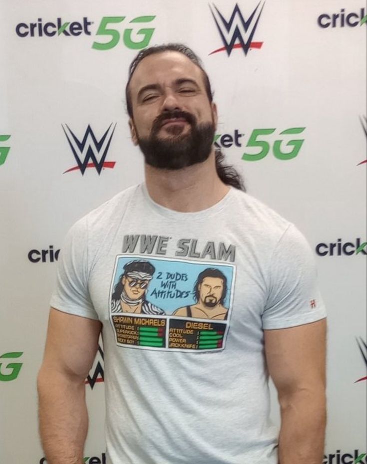 a man with long hair and beard standing in front of a white wall wearing a wrestling t - shirt