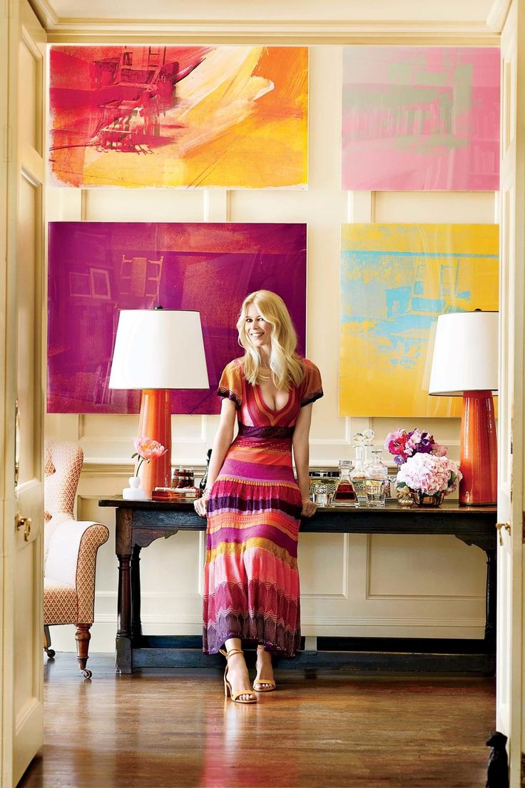 a woman standing in front of a table with two lamps and paintings on the wall behind her